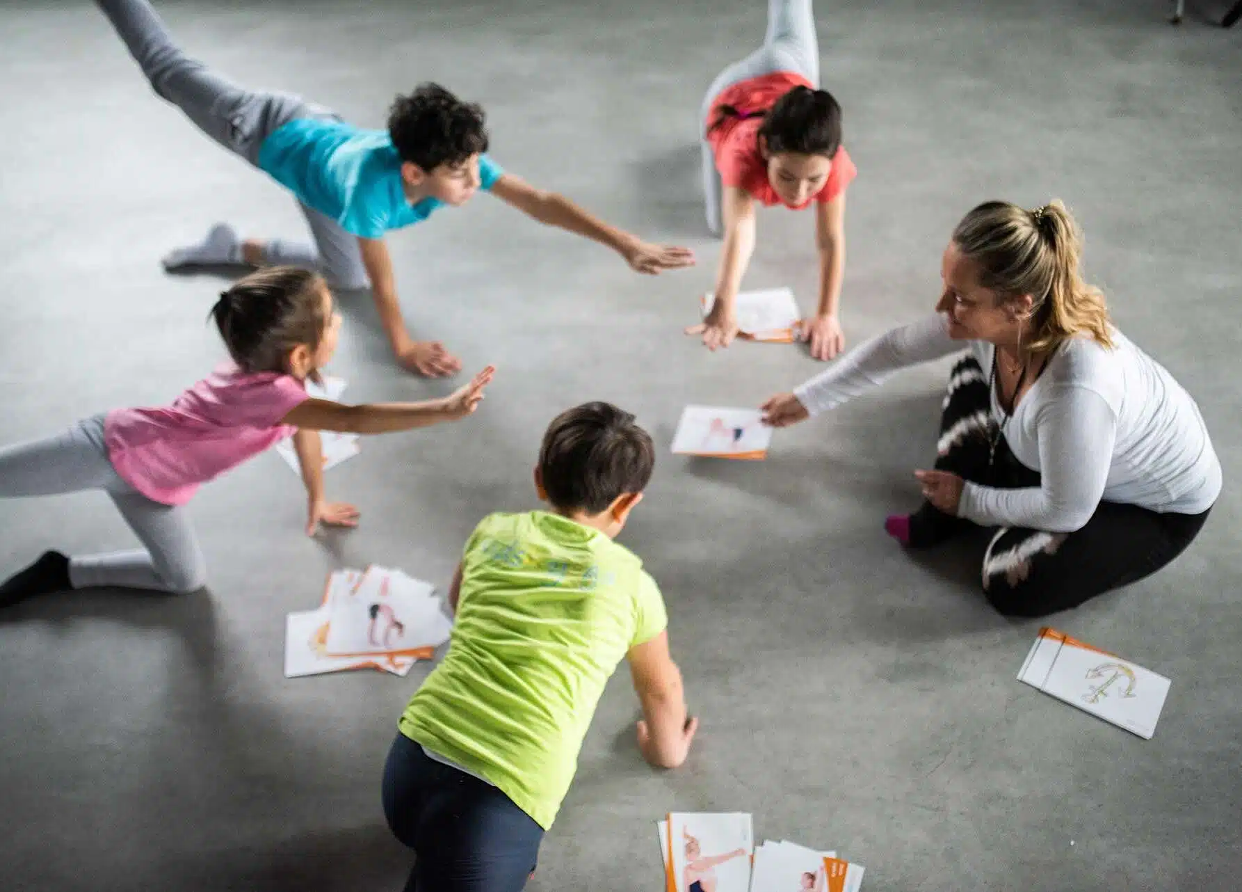 Kinder machen gemeinsam mit einer Lehrerin spielerische Yogaübungen und nutzen dabei Bildkarten als Anleitung.
