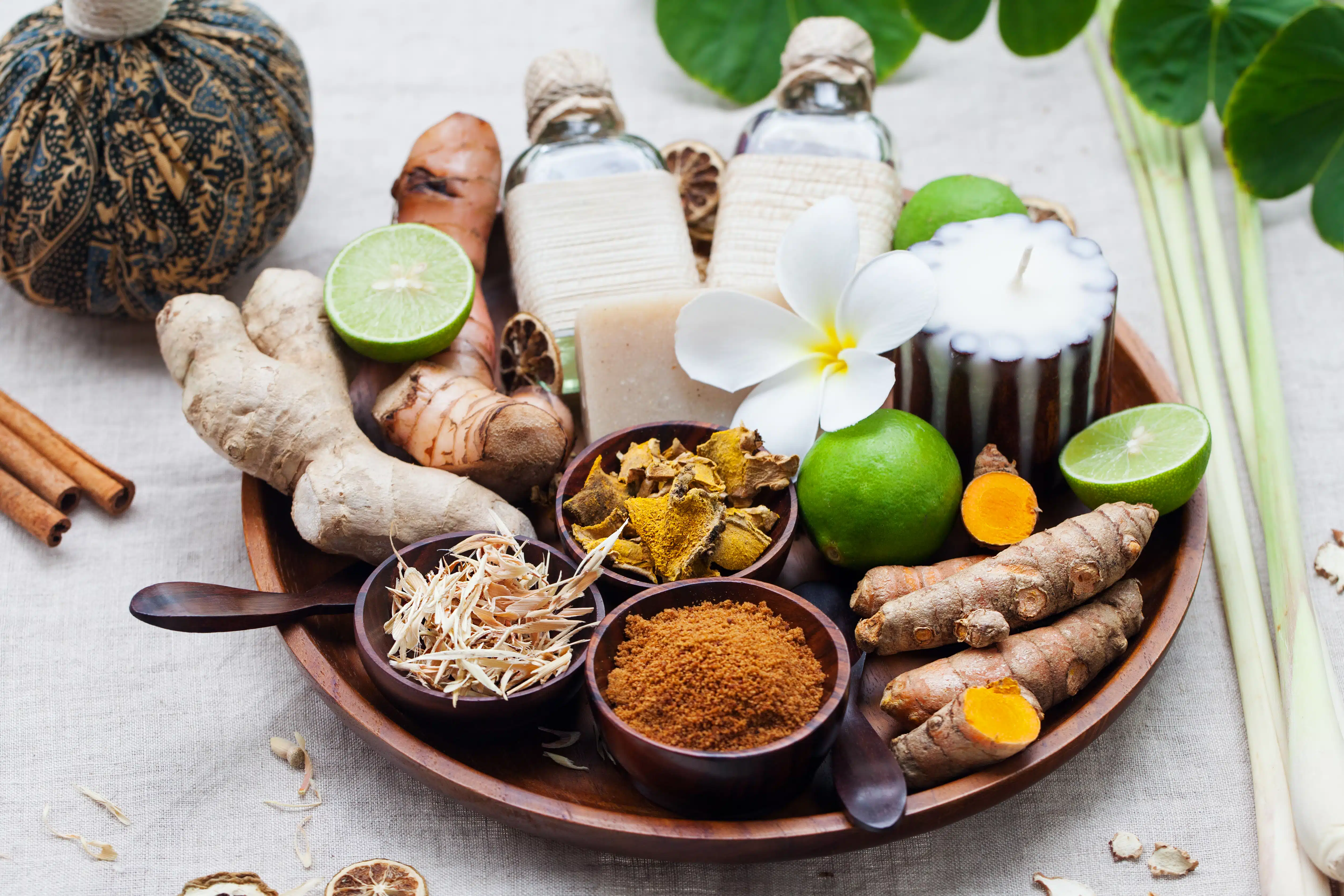 Ein Holztablett mit ayurvedischen Zutaten wie Ingwer, Kurkuma, Limetten, Zimt, Zitronengras, Kräutern, Ölen, Seife und einer Blüte. Die natürliche Anordnung vermittelt Wellness, ganzheitliche Ernährung, Gewürzkunde und ayurvedische Selbstfürsorge.