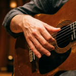 Nahaufnahme eines Händepaars, das eine klassische Gitarre mit Flamenco-Technik spielt, Finger in Position für einen Rasgueado-Anschlag, warmes Bühnen- oder Studiolicht, hölzerner Hintergrund.