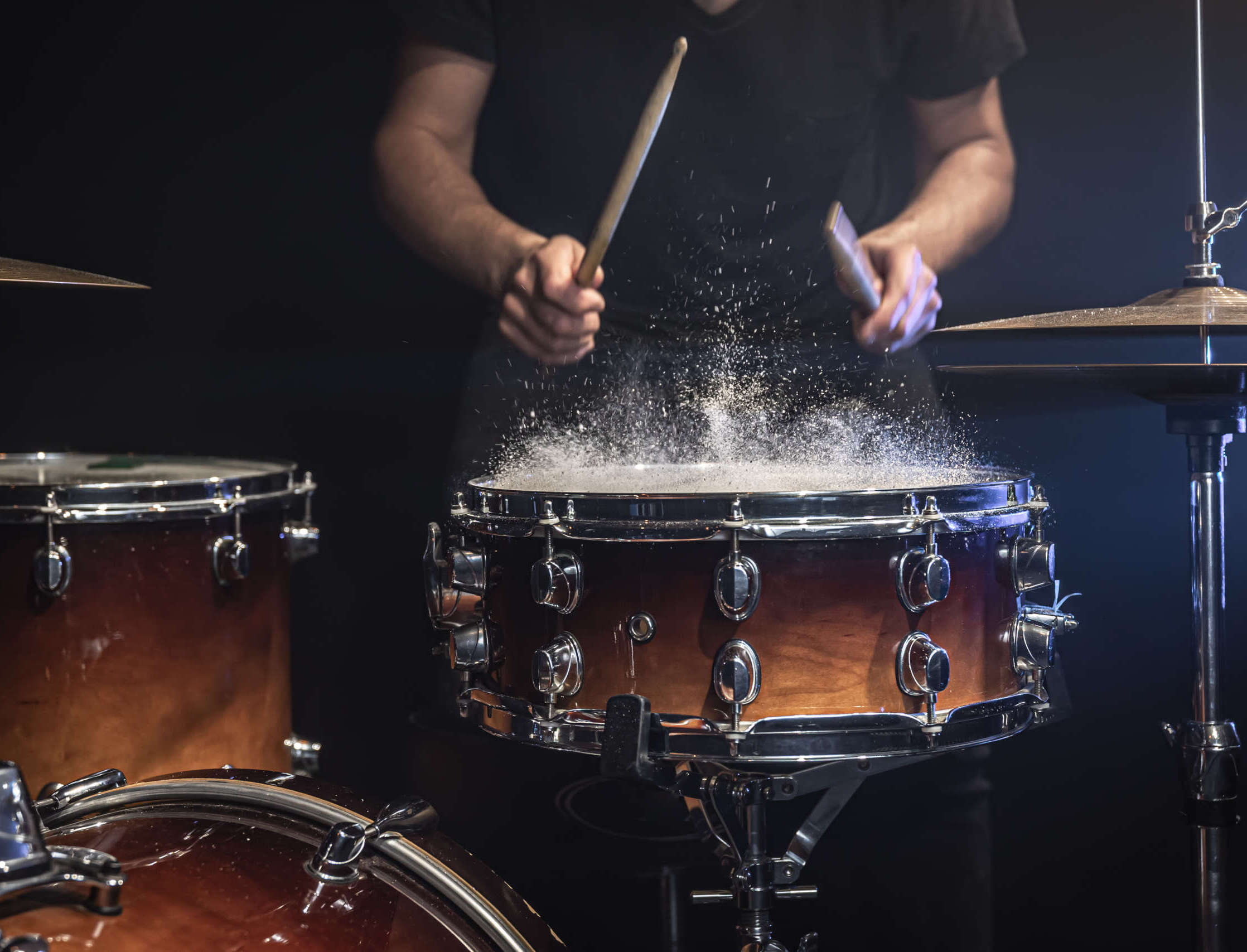 Ein Schlagzeuger spielt kraftvoll auf einer Snare-Drum, während Staub oder feine Partikel durch den Schlag sichtbar aufwirbeln. Die Szene ist dunkel und dramatisch ausgeleuchtet, mit Fokus auf Drumsticks, Trommel und Bewegung. Sie vermittelt Energie, Rhythmus, Schlagtechnik und die intensive Atmosphäre eines Schlagzeug-Onlinekurses oder Live-Trainings.