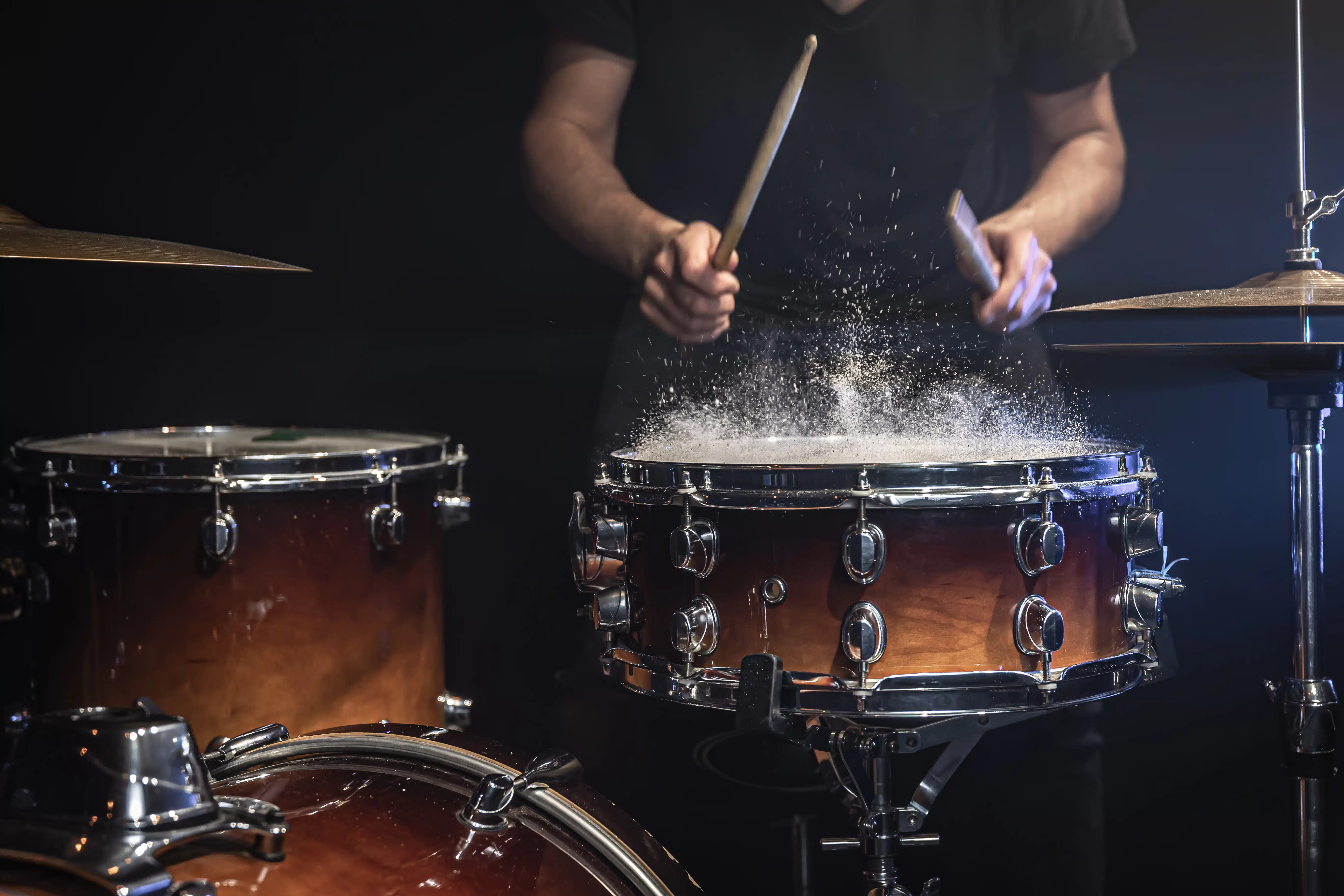 Ein Schlagzeuger spielt kraftvoll auf einer Snare-Drum, während Staub oder feine Partikel durch den Schlag sichtbar aufwirbeln. Die Szene ist dunkel und dramatisch ausgeleuchtet, mit Fokus auf Drumsticks, Trommel und Bewegung. Sie vermittelt Energie, Rhythmus, Schlagtechnik und die intensive Atmosphäre eines Schlagzeug-Onlinekurses oder Live-Trainings.