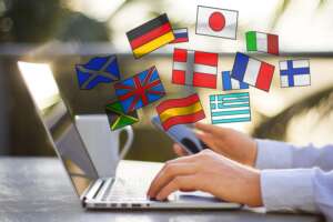 Eine Person arbeitet an einem Laptop und hält zusätzlich ein Smartphone in der Hand, während darüber verschiedene Länderflaggen als digitale Symbole eingeblendet sind. Zu sehen sind unter anderem Flaggen von Deutschland, Großbritannien, Frankreich, Italien, Spanien, Japan, Griechenland und weiteren Ländern. Die Szene vermittelt internationales Lernen, Online-Sprachkurse, Mehrsprachigkeit und digitale Kommunikation über verschiedene Kulturen hinweg.