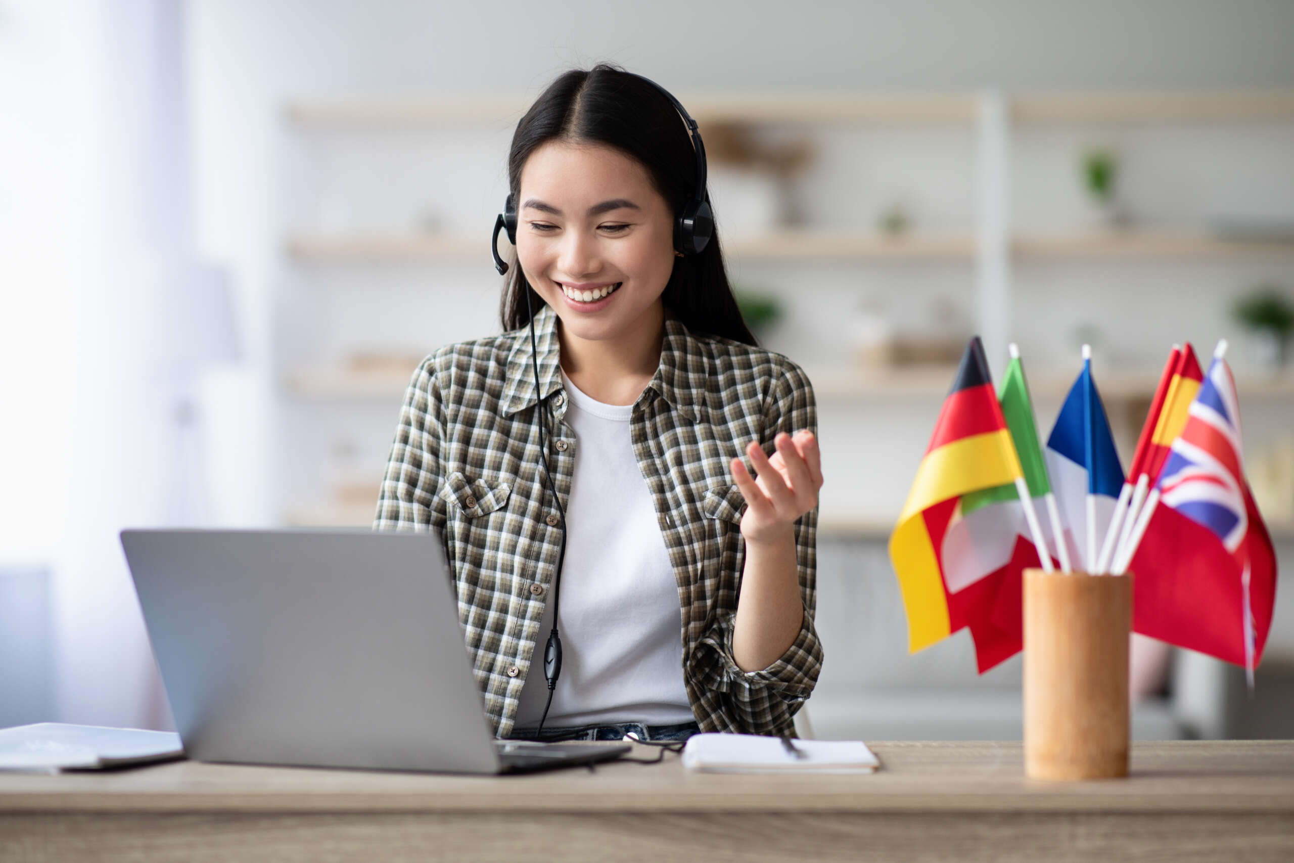 Eine lächelnde Frau sitzt mit Headset vor einem Laptop und nimmt an einem Online-Sprachkurs oder Video-Training teil. Neben ihr stehen kleine Länderflaggen, darunter Deutschland, Italien, Frankreich, Spanien und Großbritannien. Die helle, moderne Umgebung und ihr freundlicher Gesichtsausdruck vermitteln digitales Sprachenlernen, internationale Kommunikation und motiviertes Lernen von zu Hause.