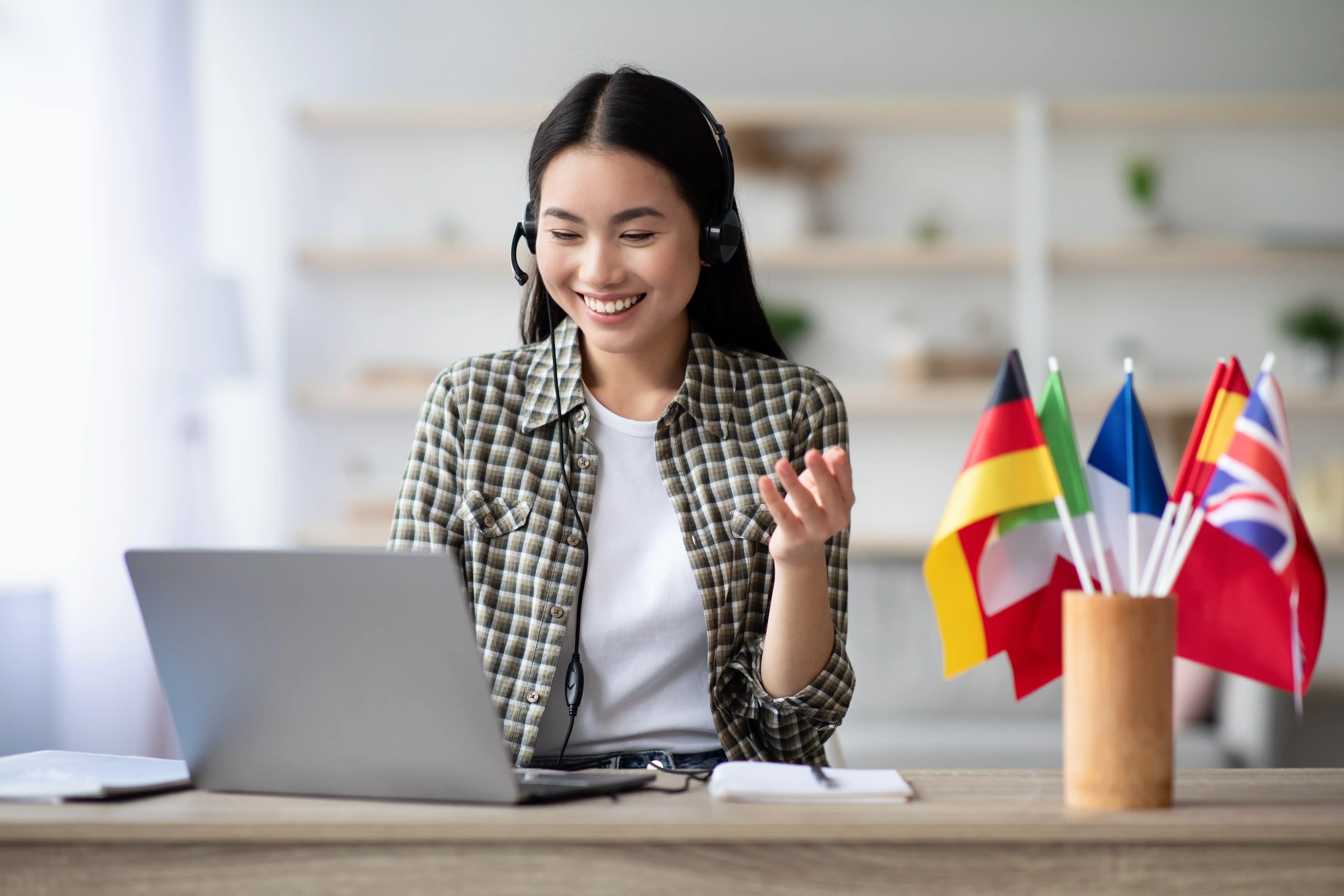 Eine lächelnde Frau sitzt mit Headset vor einem Laptop und nimmt an einem Online-Sprachkurs oder Video-Training teil. Neben ihr stehen kleine Länderflaggen, darunter Deutschland, Italien, Frankreich, Spanien und Großbritannien. Die helle, moderne Umgebung und ihr freundlicher Gesichtsausdruck vermitteln digitales Sprachenlernen, internationale Kommunikation und motiviertes Lernen von zu Hause.