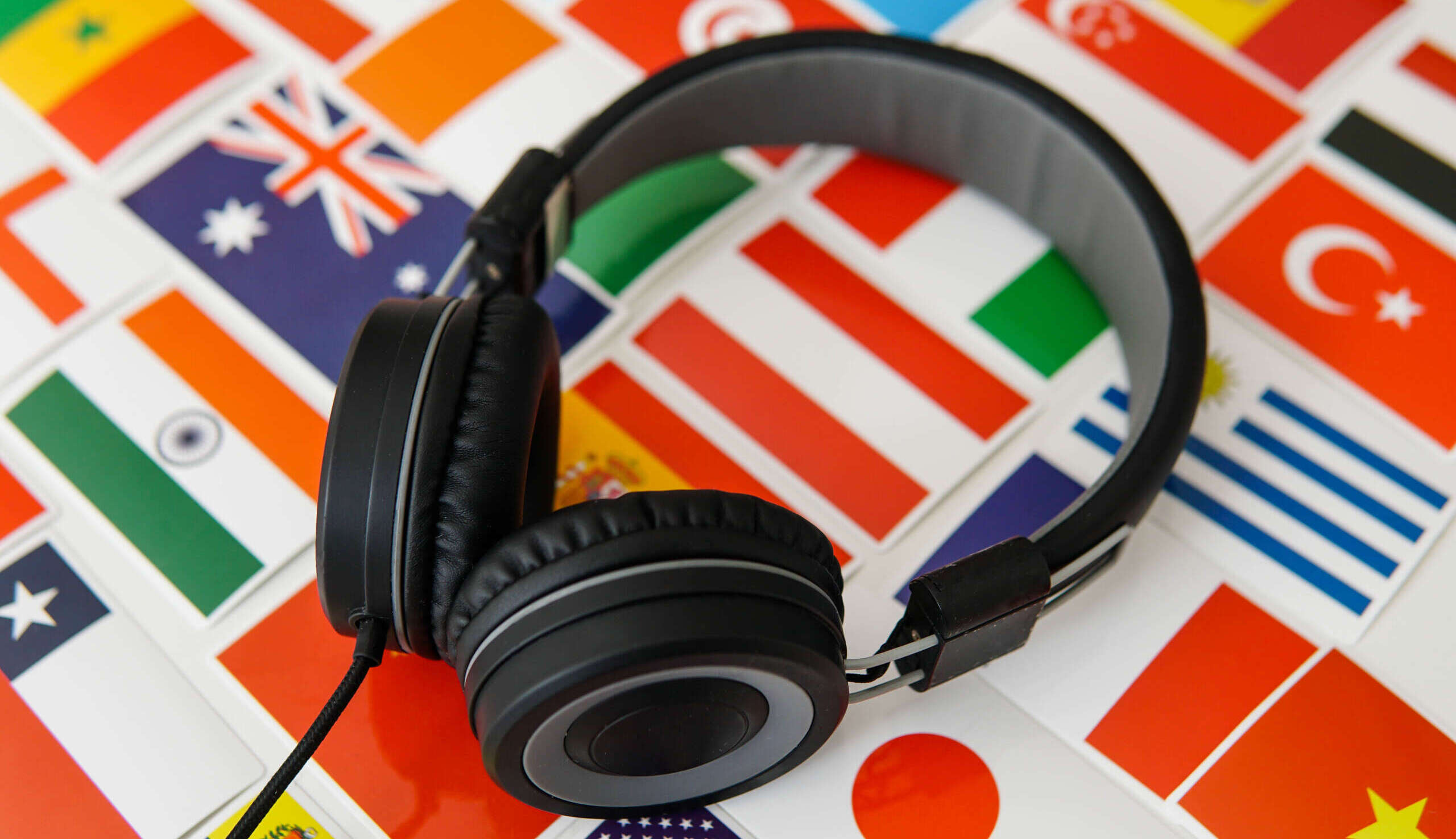Ein schwarzer Kopfhörer liegt auf einer Fläche mit vielen bunten Länderflaggen, darunter unter anderem Großbritannien, Italien, Spanien, Japan, Griechenland, die Türkei und die USA. Die Nahaufnahme verbindet Symbole internationaler Kommunikation mit dem Thema Zuhören und vermittelt eine passende Atmosphäre für Sprachenlernen, Hörverständnis, Onlinekurse und interkulturellen Austausch.