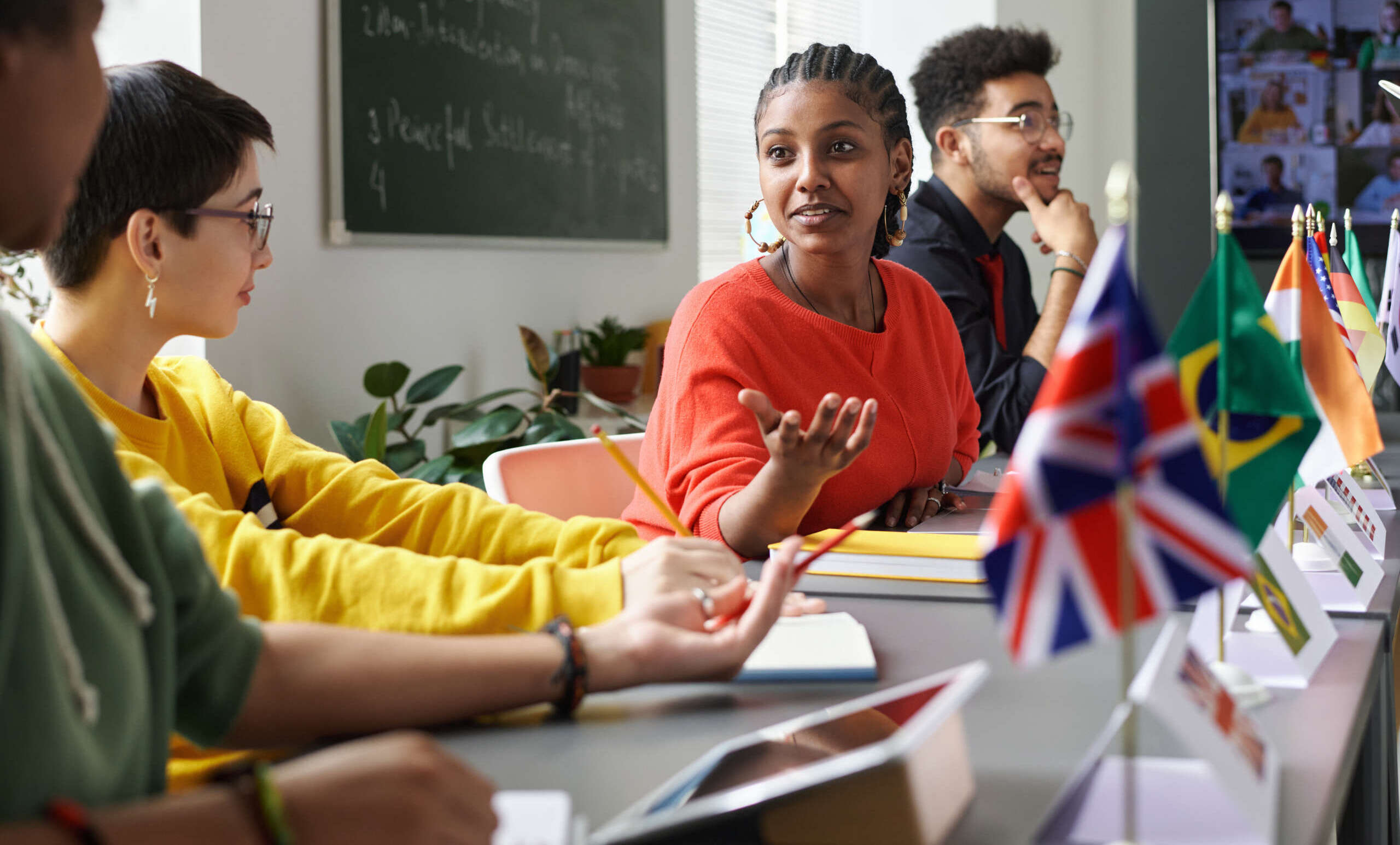 Eine Gruppe von Lernenden sitzt gemeinsam in einem hellen Sprachkursraum und diskutiert miteinander. Auf dem Tisch stehen verschiedene Länderflaggen, darunter Großbritannien und Brasilien, während im Hintergrund eine Tafel mit Notizen und ein Bildschirm mit Online-Teilnehmenden zu sehen sind. Die Szene vermittelt interkulturellen Austausch, aktives Sprachenlernen und gemeinsames Lernen in Präsenz und digitaler Verbindung.