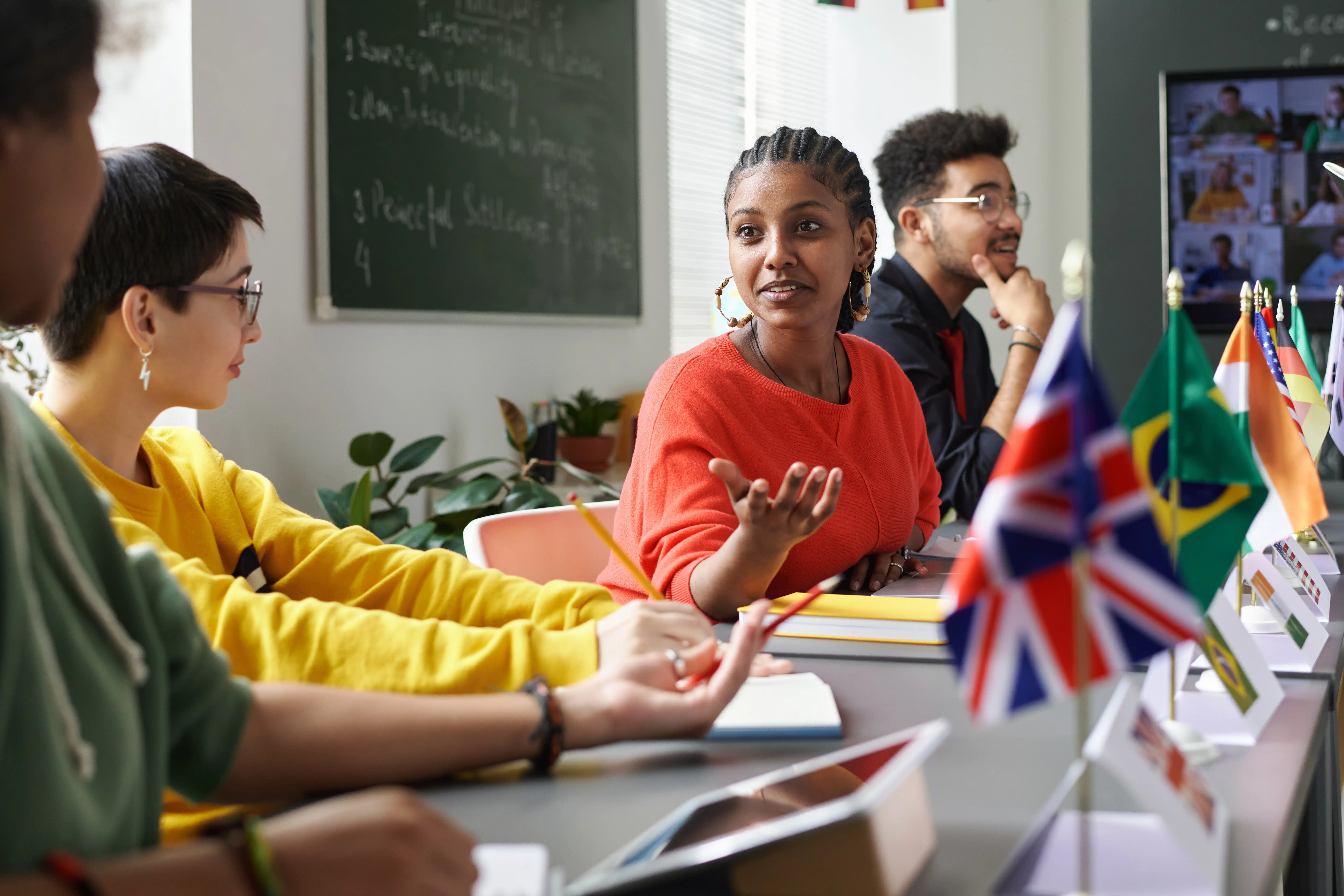 Eine Gruppe von Lernenden sitzt gemeinsam in einem hellen Sprachkursraum und diskutiert miteinander. Auf dem Tisch stehen verschiedene Länderflaggen, darunter Großbritannien und Brasilien, während im Hintergrund eine Tafel mit Notizen und ein Bildschirm mit Online-Teilnehmenden zu sehen sind. Die Szene vermittelt interkulturellen Austausch, aktives Sprachenlernen und gemeinsames Lernen in Präsenz und digitaler Verbindung.