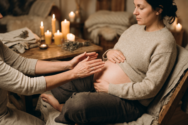 Schwangere Frau im beigen Strickpullover sitzt mit geschlossenen Augen entspannt auf einem Stuhl, während eine zweite Person sanft ihre Hände auf den freigelegten Babybauch legt. Im Hintergrund ein warmtoniges Zimmer mit mehreren brennenden Kerzen und getrockneten Kräutern auf einem Holztisch.