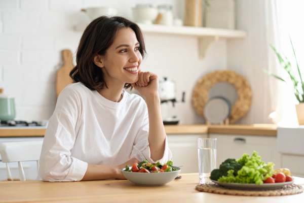 Eine lächelnde junge Frau mit dunklem, schulterlangem Haar und weißem Oversized-Shirt sitzt entspannt am Küchentisch und schaut träumerisch zur Seite. Vor ihr steht eine Schüssel mit frischem Salat, ein Glas Wasser und ein Teller mit Brokkoli, Salat und Tomaten. Im Hintergrund eine helle, modern-skandinavische Küche mit Holzakzenten und natürlichem Tageslicht.