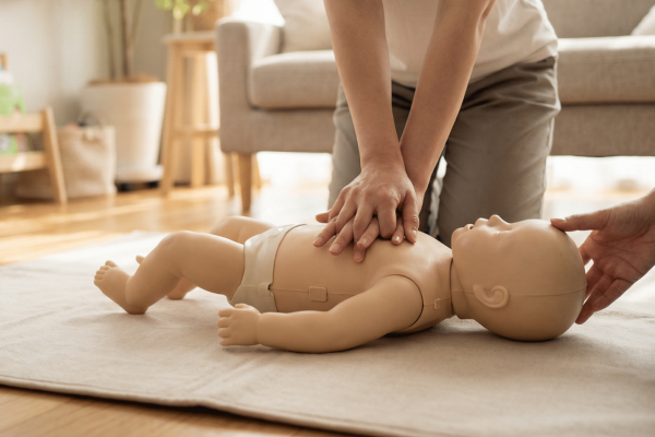 Trainingsszene mit einer Baby-Reanimationspuppe: Eine Person kniet auf dem Boden und führt Herzdruckmassage an der auf einer hellen Matte liegenden Säuglingspuppe durch, während eine zweite Person den Kopf der Puppe hält und stabilisiert. Im Hintergrund ein helles, wohnliches Zimmer mit Sofa, Zimmerpflanzen und einem Bücherregal mit Kinderbüchern.