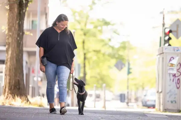 Frau in schwarzem Shirt und Jeans geht auf einem Stadtbürgersteig und schaut lächelnd auf einen schwarzen Labrador-Welpen hinunter, der mit geöffnetem Maul neben ihr läuft und zu ihr hochblickt. Im Hintergrund eine belebte Stadtstraße mit Bäumen und Ampel.