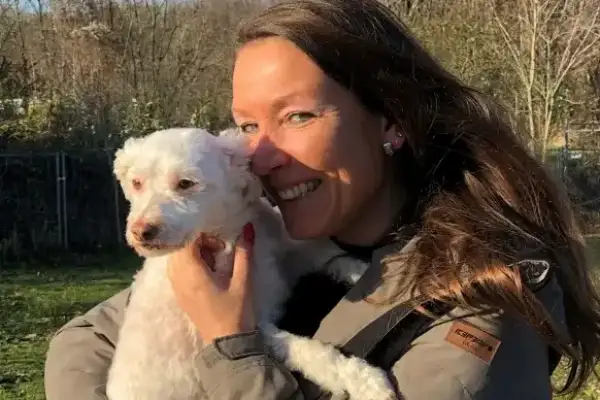 Lächelnde Frau mit langen braunen Haaren und grauer Jacke hält einen kleinen weißen Lockenhund (vermutlich Pudelmix) an ihre Wange gedrückt. Im Hintergrund eine grüne Außenanlage mit Bäumen und Zaun im Gegenlicht.