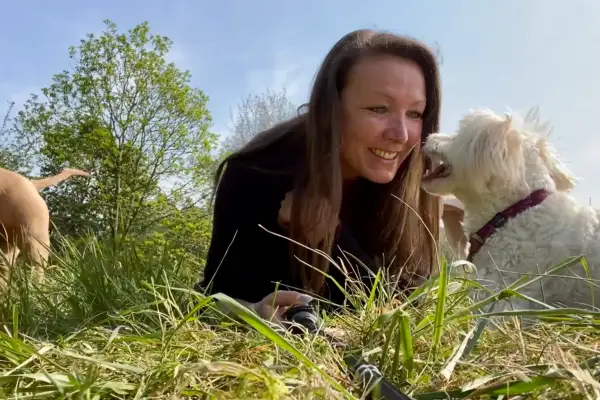 Lächelnde Frau mit langen braunen Haaren und schwarzem Oberteil liegt im Gras und lässt sich von einem weißen Lockenhund mit lila Halsband ins Gesicht lecken. Im Hintergrund ein zweiter brauner Hund sowie Büsche und blauer Himmel.