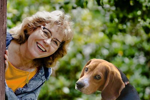 Eine herzlich lachende Frau mit kurzen, lockigen, blonden Haaren und Brille lehnt sich von links um einen Baumstamm und schaut einen Beagle mit schwarz-braunem Fell an. Der Hund sitzt ruhig daneben und blickt aufmerksam nach vorne. Im Hintergrund üppiges grünes Laub im Sonnenlicht.