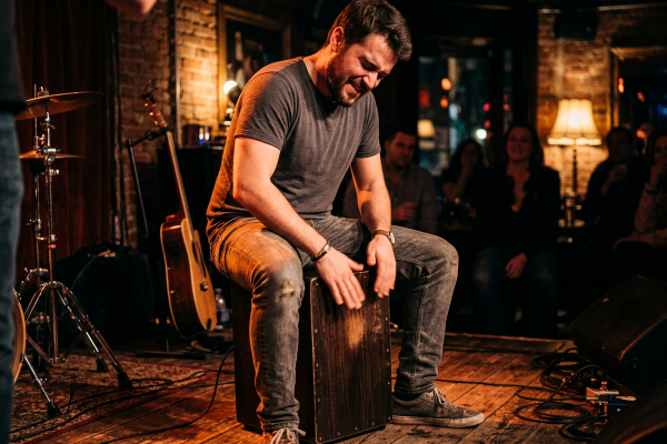 Ein Mann sitzt auf einer kleinen Bühne auf einer Cajón und spielt konzentriert mit beiden Händen darauf. Warmes Bühnenlicht, Backsteinwände, Holzboden, Kabel, ein Mikrofon, ein Schlagzeugständer und eine Gitarre im Hintergrund schaffen eine intime Live-Atmosphäre. Im Publikum sitzen mehrere Personen, die dem Auftritt aufmerksam folgen. Die Szene vermittelt Rhythmusgefühl, Percussion, musikalische Energie und das Spielen einer Cajón in einem kleinen Konzert- oder Kursumfeld.