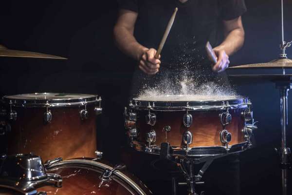 Ein Schlagzeuger spielt kraftvoll auf einer Snare-Drum, während Staub oder feine Partikel durch den Schlag sichtbar aufwirbeln. Die Szene ist dunkel und dramatisch ausgeleuchtet, mit Fokus auf Drumsticks, Trommel und Bewegung. Sie vermittelt Energie, Rhythmus, Schlagtechnik und die intensive Atmosphäre eines Schlagzeug-Onlinekurses oder Live-Trainings.