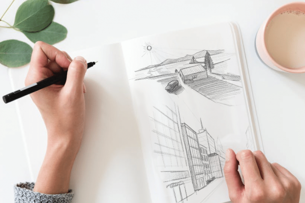 Draufsicht auf zwei Hände, die ein aufgeschlagenes Skizzenbuch halten und mit einem schwarzen Fineliner zeichnen. Im Skizzenbuch sind zwei Bleistiftzeichnungen zu sehen: eine Landschaft mit Haus, Baum und Bergen, sowie ein perspektivisches Stadtgebäude. Daneben auf dem weißen Tisch eine rosafarbene Kaffeetasse und ein Eukalyptuszweig.