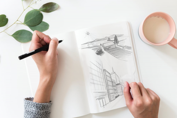 Draufsicht auf zwei Hände, die ein aufgeschlagenes Skizzenbuch halten und mit einem schwarzen Fineliner zeichnen. Im Skizzenbuch sind zwei Bleistiftzeichnungen zu sehen: eine Landschaft mit Haus, Baum und Bergen, sowie ein perspektivisches Stadtgebäude. Daneben auf dem weißen Tisch eine rosafarbene Kaffeetasse und ein Eukalyptuszweig.