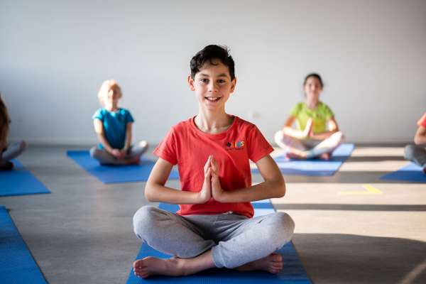 Lächelnder Junge im roten T-Shirt sitzt im Schneidersitz auf einer blauen Yogamatte mit Händen in Namaste-Haltung. Im Hintergrund weitere Kinder auf blauen Matten in einem hellen Yogaraum.